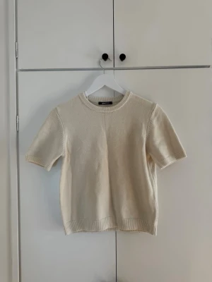 Kortärmad stickad tröja i beige - En stilren, kortärmad stickad tröja i mjuk beige färg. Tröjan har rund halsringning och ribbade kanter vid ärmslut och nederkant. Perfekt för lager på lager och enkel att matcha med olika outfits.