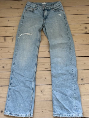 Ljusblå raka jeans med slitning - Säljer ett par ljusblå jeans med rak passform och hög midja. Jeansen har en sliten detalj på vänster lår och klassiska fem fickor. Materialet är denim i bomull och de har en vintagekänsla. Perfekta för en avslappnad look.
