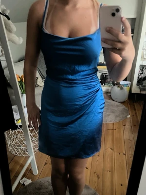 Blå kort satinklänning med smala band - Säljer en snygg blå kort klänning i satin med smala axelband och draperad urringning. Klänningen har en figurnära passform och är perfekt för dig som vill ha en stilren och trendig look.