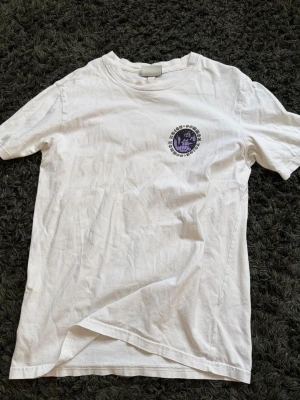 Vit t-shirt med tryck Common Noise - Vit t-shirt med svart och lila tryck på bröstet och ryggen där det står 'Common Noise Common Voice' i cirkelform. T-shirten har klassisk passform och rund hals. Perfekt för dig som gillar grafiska prints och enkel stil.
