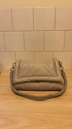Beige vadderad handväska med flätat handtag - Säljer en beige vadderad handväska med ett snyggt flätat handtag. Väskan har magnetknäppning och en mjuk, puffig design. Perfekt för dig som gillar stilrena accessoarer med en twist. Passar till det mesta och är enkel att matcha. Den är synligt använd inuti men inget man lägger märke till. 