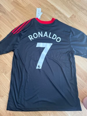 Manchester United Ronaldo tröja svart - Säljer en svart Manchester United matchtröja från Adidas med Ronaldo och nummer 7 på ryggen. Tröjan har röda detaljer på axlarna, klubbmärke och Adidas-logga på bröstet samt Tezos sponsortryck. Tillverkad i lätt och ventilerande polyester.