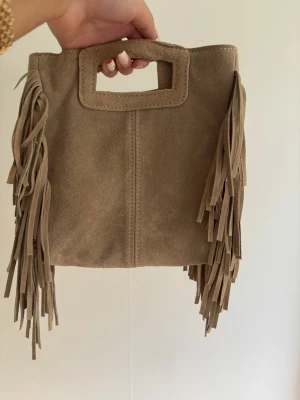 Maje liknande väska i mocka - Cool beige handväska i mocka med rektangulärt handtag och långa fransar på sidorna. Väskan har ett innerfack med dragkedja och avtagbar axelrem. Perfekt för dig som gillar boho-stil och vill sticka ut med accessoarer.