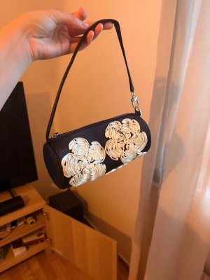 Svart cylinderformad handväska med blommor - Cool svart handväska i cylinderform med dekorativa vita blommor på framsidan. Väskan har ett smalt axelband och dragkedja upptill. Materialet ser ut att vara konstläder och detaljerna är i vitt för en snygg kontrast. Från caia 😍 perfekt storlek och väldigt fin och unik 💕aldrig Andväd ☹️pris kan diskuteras 👌