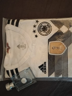 Tysklands matchtröja Adidas herr - Säljer en vit och svart Tyskland matchtröja från Adidas med officiella märken, fyra stjärnor och FIFA-märke på bröstet. Tröjan har klassiska svarta ränder och rund hals. Perfekt för fotbollsfans som vill ha en snygg och bekväm supportertröja. Strl 146