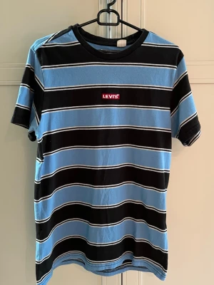 Randig blå och svart Levi's t-shirt - Säljer en snygg randig t-shirt från Levi's i blått och svart med vita detaljer. T-shirten har relaxerad passform, korta ärmar och rund hals. Levi's-loggan sitter broderad på bröstet. Perfekt till jeans eller shorts.