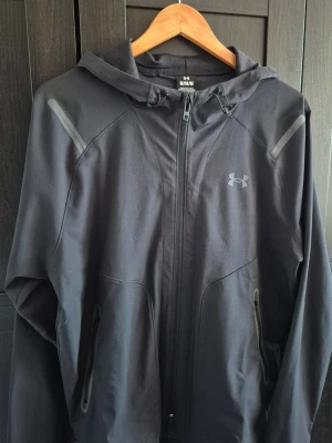 Svart hoodie från Under Armour - Snygg svart hoodie från Under Armour med dragkedja framtill, två fickor med dragkedja och diskret logga på bröstet. Tillverkad i ett mjukt och stretchigt material. Huva med dragsko för extra stil och funktion. Perfekt för dig som gillar sportig och clean look.