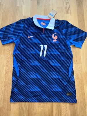 Blå Frankrike matchtröja Nike olise  - Snygg blå matchtröja från Nike med Frankrikes landslagsemblem, fotbollspelaren Olise och nummer 11 på både fram- och baksidan. Tröjan har korta ärmar, vit krage och röd detalj vid halsen. Materialet är lätt och andas, perfekt för fotboll eller träning.