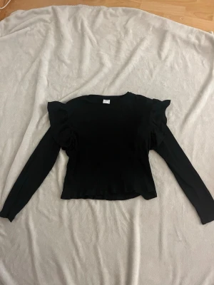 Svart långärmad topp med volang från Zara - Snygg svart långärmad topp med volangdetaljer vid axlarna och ribbad struktur. Toppen har en figurnära passform.