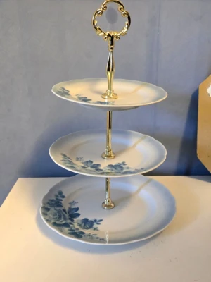  Fyrklövern, Julia Våningsfat i porslin med blommönster - Snyggt våningsfat i tre plan med vita porslinstallrikar och blått blommönster. Fatet har en dekorativ guldfärgad metallstång med handtag upptill, perfekt för kakor eller små bakverk. Klassisk och stilren design som passar till fika eller kalas.