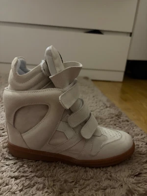 Isabell marant skor - Snygga beiga sneakers med kilklack och kardborreband. Skorna har en hög siluett och är gjorda i mockaliknande material med gummisula i brunt. Perfekta för dig som vill ha lite extra höjd och en unik look. Köpt dessa på plick , är ganska säker på att de är kopior då jag inte fick något äkthetsbevis och säljer då de ej kmr till användning❤️storlek 38 men passar även 39. Pris kan diskuteras vid snabb affär 💕