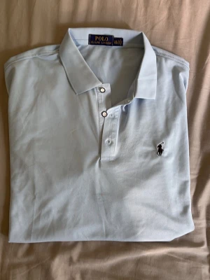 Ljusblå piké från Polo Ralph Lauren - Säljer en klassisk ljusblå pikétröja från Polo Ralph Lauren med korta ärmar och knappslå framtill. Tröjan har den ikoniska broderade loggan på bröstet och är tillverkad i mjuk bomull. Perfekt för en clean och stilren look.