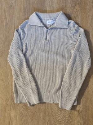 Beige stickad tröja med dragkedja - Beige stickad tröja från East West med halv dragkedja framtill och bred krage. Tröjan har lång ärm och ribbad struktur som ger en avslappnad men ändå stilren look. Perfekt till jeans eller för en enkel och snygg outfit.