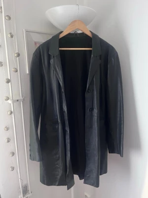 Svart lång skinnjacka med knappar - Svart skinnkappa i längre modell med klassisk krage och knäppning framtill. Jackan har två fickor och en rak passform. 
