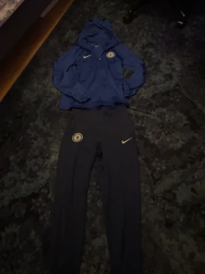 Chelsea Nike blå träningsset -  Passar xs oxå Säljer ett blått träningsset från Nike med Chelsea FC-logga. Setet består av en hoodie med huva och dragkedja samt matchande byxor med resår i midjan. Perfekt för träning eller chill. Båda plaggen har Nike-logga och Chelsea-märke på bröst och ben.