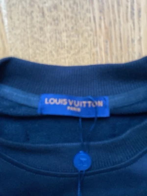 Svart sweatshirt från Louis Vuitton - Snygg svart sweatshirt från Louis Vuitton med klassisk rund hals och ribbade muddar. På bröstet finns ett stort LV-logotyptryck i blått och gult. Perfekt för dig som gillar exklusiva streetwear-plagg.