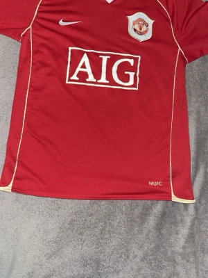 Manchester United matchtröja röd Nike - Klassisk röd Manchester United matchtröja från Nike med vitt AIG-tryck på bröstet och klubbmärke på vänster sida. Tröjan har vita detaljer längs sidorna och V-ringad hals. Perfekt för fotbollsfans och samlare.