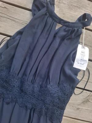 Mörkblå långklänning med spetsdetalj - Elegant mörkblå långklänning med vackra spetsdetaljer i midjan och hög halsringning. Klänningen har ärmlös design. St 38