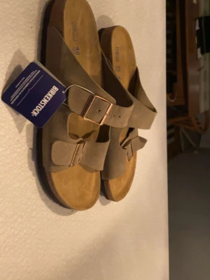  Birkenstock sandaler - Snygga beige sandaler från Birkenstock med två breda remmar i mocka och justerbara metallspännen. Fotbädden är i kork och sulan är i gummi för extra komfort. Perfekta för en avslappnad och trendig stil.