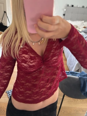 Vinröd spetsblus med v-ringning - Säljer en vinröd, transparent spetsblus med v-ringning och lång ärm. Toppen har en snygg twistdetalj framtill och är croppad i modellen. Perfekt för dig som vill ha en trendig och feminin look.