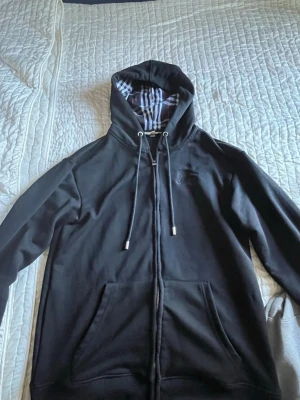 Svart hoodie från Burberry med dragkedja - Svart hoodie från Burberry med klassiskt rutigt foder i huvan och dragsko. Tröjan har dragkedja framtill och två fickor. Perfekt för en stilren och avslappnad look. Materialet känns mjukt och bekvämt.