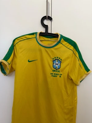 Brasilien fotbollströja R. Carlos - Brasilien fotbollströja från Nike med gröna detaljer och tryck. På bröstet finns CBF-emblemet och texten 'FIFA World Cup France 98'. På ryggen står det 'R. CARLOS' och nummer 6. Perfekt för dig som älskar fotboll och klassiska landslagströjor. Storlek M men kan också passa S, då det är en lite mindre passform.