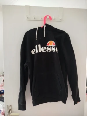 Svart Ellesse hoodie med logga - Snygg svart hoodie från Ellesse med stor vit logga och orange detalj på bröstet. Vita dragsnören i huvan. Perfekt för en chill och sportig look. Tillverkad i mjukt material för extra komfort. Har tyvärr gått sönder vid ena armen men inget man inte kan fixa.