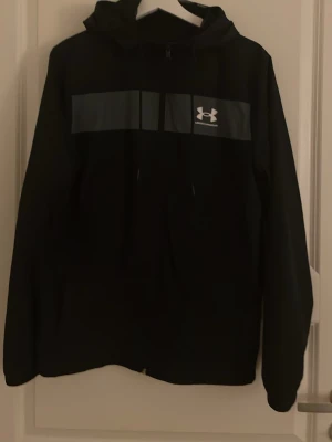 Svart hoodie från Under Armour - Svart hoodie från Under Armour med huva och dragsko. På bröstet finns en bred grå panel med Under Armour-loggan i vitt. Hoodien har långa ärmar och en klassisk passform. Perfekt för en sportig och avslappnad stil.