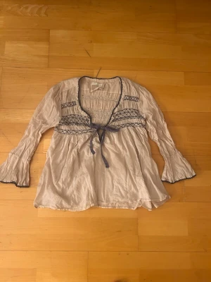 Beige blus med broderi och knytband - Söt beige blus med långa ärmar, mörkblå broderade detaljer och volangkant vid ärmslut. Blusen har ett knytband under bysten och lätt rynkade partier för en bohemisk look. Tillverkad i ett tunt, luftigt tyg som känns skönt mot huden. (Se bild 2 för en liten reva i armen)