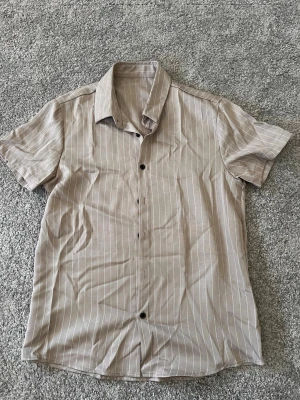 Beige randig kortärmad skjorta - Snygg beige skjorta med vita ränder och korta ärmar. Skjortan har klassisk krage och knäppning framtill. Perfekt för en avslappnad och stilren look.