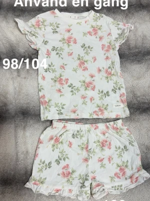 Blommig pyjamas med volangdetaljer - Tvådelad pyjamas med t-shirt och shorts i vitt tyg med rosa och gröna blommor. T-shirten har volangärmar och shortsen har elastisk midja samt volangkant nedtill. Perfekt för mysiga kvällar och nätter.🌸 Använd en gång. Storlek 98/104