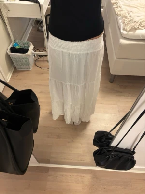 Vit lång kjol med smock i midjan - Lång, luftig vit kjol med bred smock i midjan och volangdetaljer. Kjolen har en avslappnad passform och är tillverkad i ett lätt tyg som passar perfekt till varmare dagar. Enkel att matcha med olika toppar för en fräsch look.