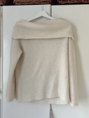 Off shoulder stickad tröja cremevit - Säljer en cremevit stickad tröja med off shoulder-design och bred krage. Tröjan har lång ärm och är ribbstickad för en mysig och trendig look. Perfekt för dig som gillar stilrena och bekväma plagg.