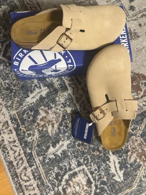 Birkenstock boston bs ’sand’ - Säljer ett par beige Birkenstock Boston tofflor i mocka med justerbart spänne i metall. Klassisk slip-in modell med mjuk fotbädd och stilren design. Perfekta för dig som vill ha både komfort och trendig look.