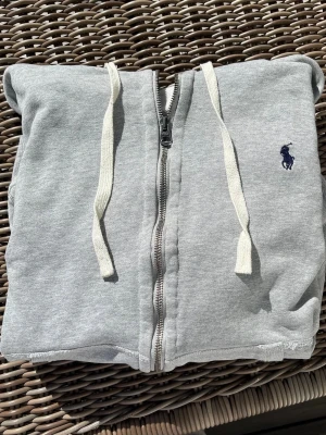 Grå zip hoodie från Polo Ralph Lauren - Snygg grå hoodie med dragkedja från Polo Ralph Lauren. Klassisk design med vit dragsko i huvan och mörkblå logga broderad på bröstet. Tillverkad i mjuk bomullsblandning, perfekt för chill dagar. Ribbstickade muddar vid ärmar och nederkant. Det är barn storlek men det är som xs i vuxen 100%.