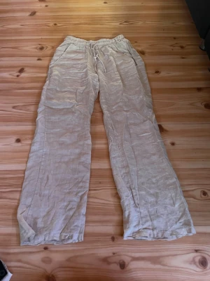 Beiga vida linnebyxor - Snygga och luftiga linnebyxor i beige med raka, vida ben och resår i midjan. Perfekta för en avslappnad och stilren look. Byxorna har fickor på sidorna och är tillverkade i ett mjukt linnematerial.