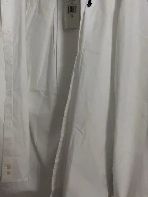 Vit slim fit skjorta från Polo Ralph Lauren (Äkta) - Klassisk vit skjorta från Polo Ralph Lauren i slim fit-modell. Skjortan har långa ärmar, knappar framtill och den ikoniska broderade loggan på bröstet. Tillverkad i mjuk bomull som känns skön mot huden. Perfekt för en clean och stilren look.