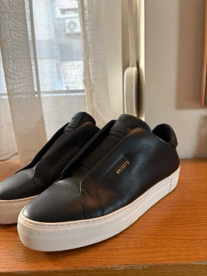 Svarta sneakers från Axel Arigato - Stilrena svarta sneakers från Axel Arigato i slätt läder med vit tjock sula. Diskret guldtext på sidan och minimalistisk design utan synliga snören. Perfekta för dig som gillar clean och modern look.