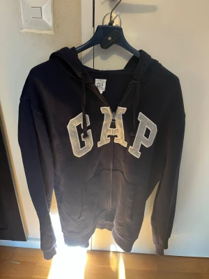 Mörkblå GAP zip hoodie med huva - Mörkblå hoodie från GAP med dragkedja framtill och stor logga i vitt på bröstet. Utrustad med huva och snören samt två fickor framtill. Perfekt för en avslappnad och cool stil.