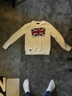 Vit stickad tröja Morris R. Club - Vit stickad tröja från Morris R. Club med brittiska flaggan i rött, svart och vitt på bröstet. Tröjan har rund halsringning och ribbade muddar vid ärmslut och nederkant. Perfekt för dig som gillar klassisk och stilren design.
