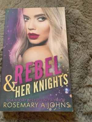 Rebel & Her Knights bok - En färgstark pocketbok med titeln 'Rebel & Her Knights' av Rosemary A Johns. Omslaget har en porträttbild av en kvinna med blont hår och starka färger i lila och guld. Perfekt för dig som gillar romantisk och dramatisk skönlitteratur.
