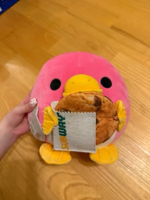 Zuru Snackles Subway plyschpingvin - Söt plyschpingvin från Zuru Snackles Series 2, rosa med gula fötter och näbb. Pingvinen håller en Subway-kaka i en tygpåse med Subway-logga. Perfekt som samlarleksak eller dekoration.