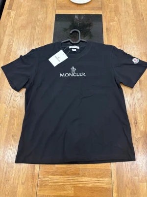 Svart Moncler t-shirt - Snygg svart t-shirt från Moncler med vit broderad logga på bröstet och en patch på ärmen. Klassisk rund halsringning och korta ärmar. Tillverkad i mjuk bomull som känns skön mot huden. Perfekt för dig som vill ha en stilren och trendig look.