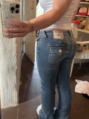 Blå bootcut jeans från True Religion - vingar  - Säljer ett par blå lågmidjade bootcut jeans från True Religion med ikoniska kontrastsömmar och detaljerade bakfickor med lock och knapp. Jeansen har en klassisk femficksmodell och patch med logga bak i midjan. Tillverkade i denim med lätt stretch för skön passform. De har vingar på baksidan samt är uppsydda till någon som är runt 160cm☺️ Använda kanske 3 gånger!