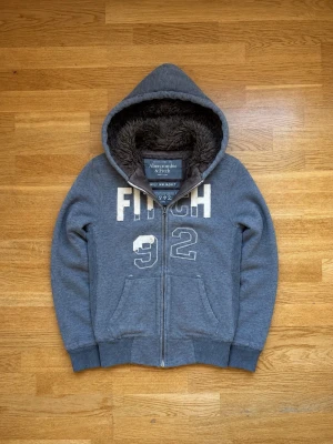 Grå Abercrombie & Fitch Wolf Jacket med innerfoder - Mysig grå hoodie från Abercrombie & Fitch med dragkedja, stora fickor och broderad text 'FITCH 92' på bröstet. Huvan är fodrad med mjuk, brun fuskpäls för extra komfort och värme. Perfekt för chill dagar och streetwear-stil.