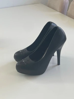 Svarta pumps med dekorationer  - Snygga svarta pumps med hög klack och rundad tå. Skorna har dekorativa stenar framtill som ger en extra touch. Tillverkade i ett material som liknar konstläder och har en klassisk siluett som passar till många olika stilar.