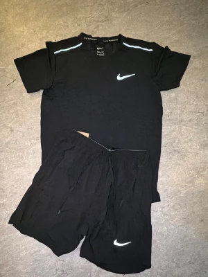 Svart träningsset från Nike - Säljer ett svart träningsset från Nike med t-shirt och shorts. T-shirten har reflekterande detaljer på axlarna och Nike-logga på bröstet. Shortsen har även de en reflekterande Nike-logga. Perfekt för löpning eller gym. Materialet är lätt och andas.