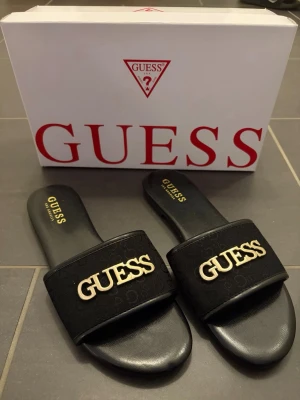 Svarta sandaler från Guess - Stilrena svarta sandaler från Guess med bred rem i textil och guldigt Guess-logotyp framtill. Aldrig använda 