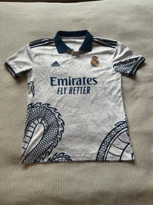 Real Madrid draktröja Adidas - Säljer en vit Real Madrid matchtröja från Adidas med mörkblå detaljer och ett stort drakmönster över hela tröjan. Tröjan har korta ärmar, pikékrage och klubbmärke på bröstet. Materialet är lätt och andas, perfekt för fotboll eller träning.