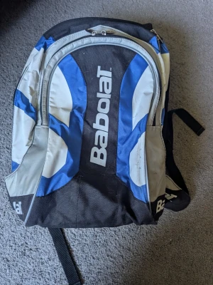 Babolat svart och blå tennis ryggsäck  - Ryggsäck från Babolat i svart, blått och vitt med sportig design. Stort huvudfack, flera dragkedjeförsedda fickor och vadderade axelband. Perfekt för träning eller skola, tillverkad i slitstark syntet. Tydlig Babolat-logga på både fram- och sidopanel.